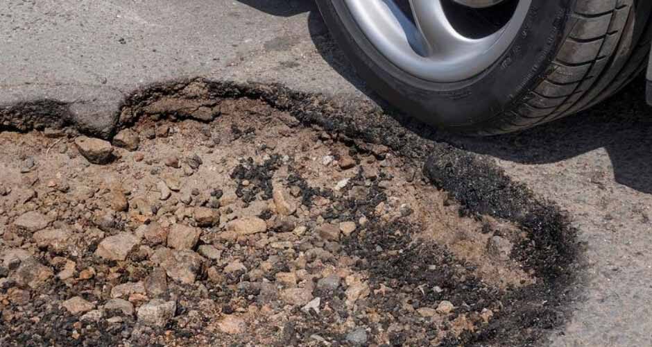 A car wheel is shown next to a large pothole on an asphalt road